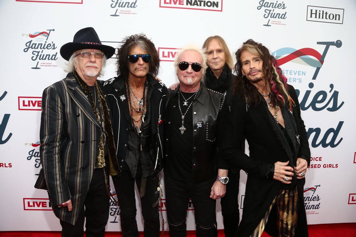 The members of Aerosmith stand in a line in front of a step and repeat banner.
