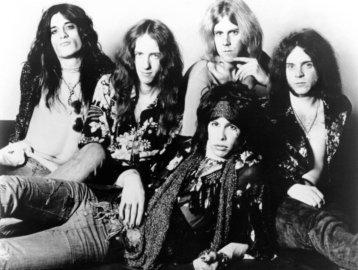 A black and white picture of the members of Aerosmith posing together. Steven Tyler lays in front of his bandmates.