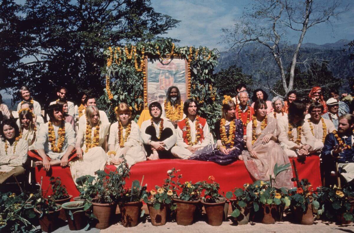 The Beatles sit with a group on a red table while in India. There are flowers in front of them.