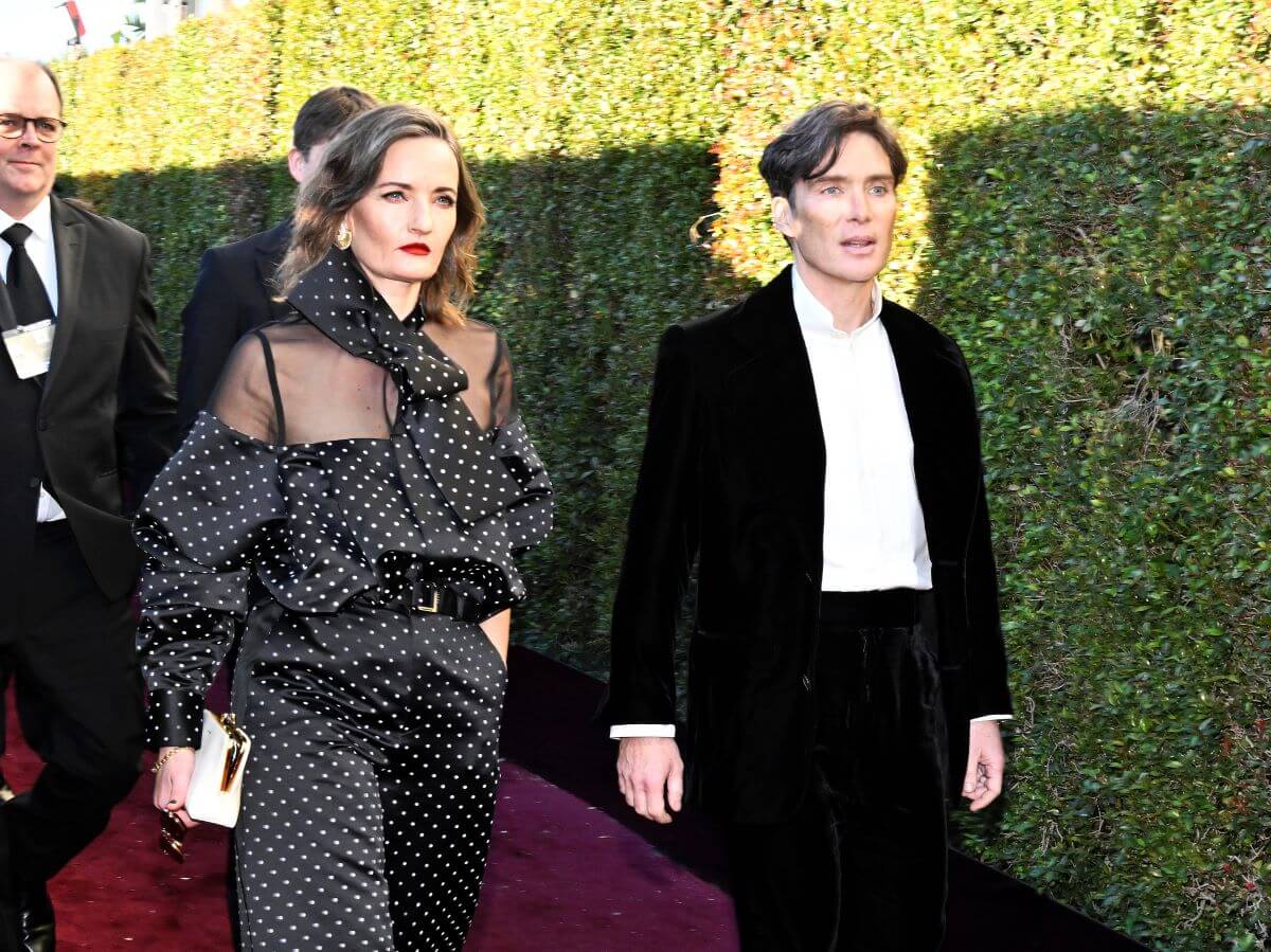 Cillian Murphy and his wife Yvonne McGuinness walk on a red carpet together next to bushes. They both wear black.