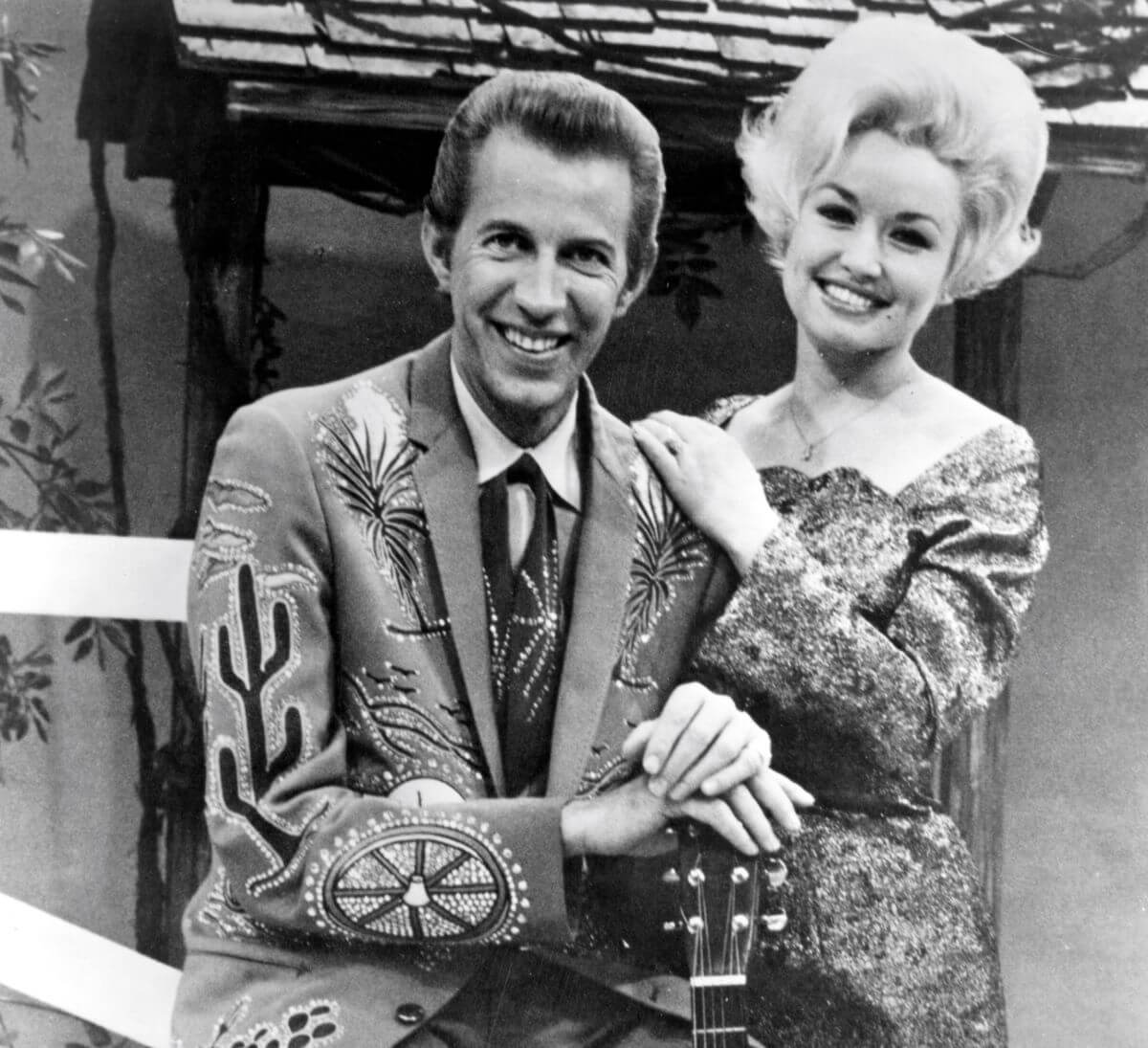 A black and white picture of Dolly Parton resting her hands on Porter Wagoner's shoulders. He rests his hands on a guitar.