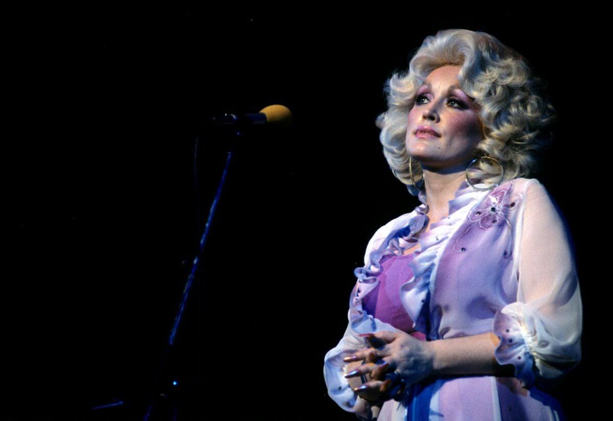 Dolly Parton wears a purple dress and holds a microphone.