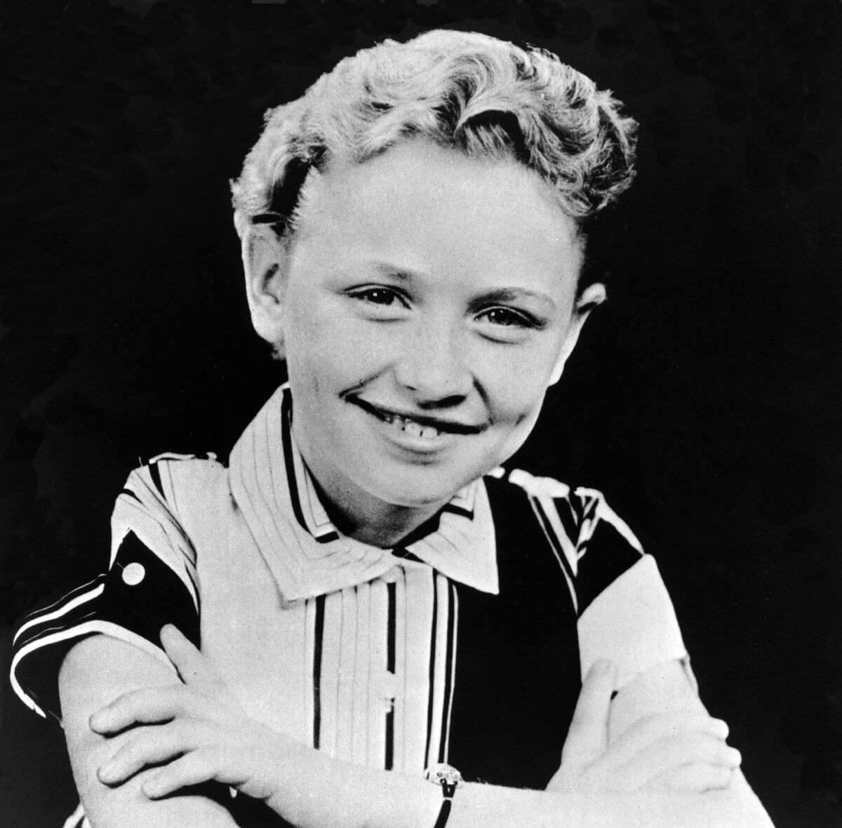 A black and white picture of a young Dolly Parton wearing a collared shirt. She sits with her arms crossed.