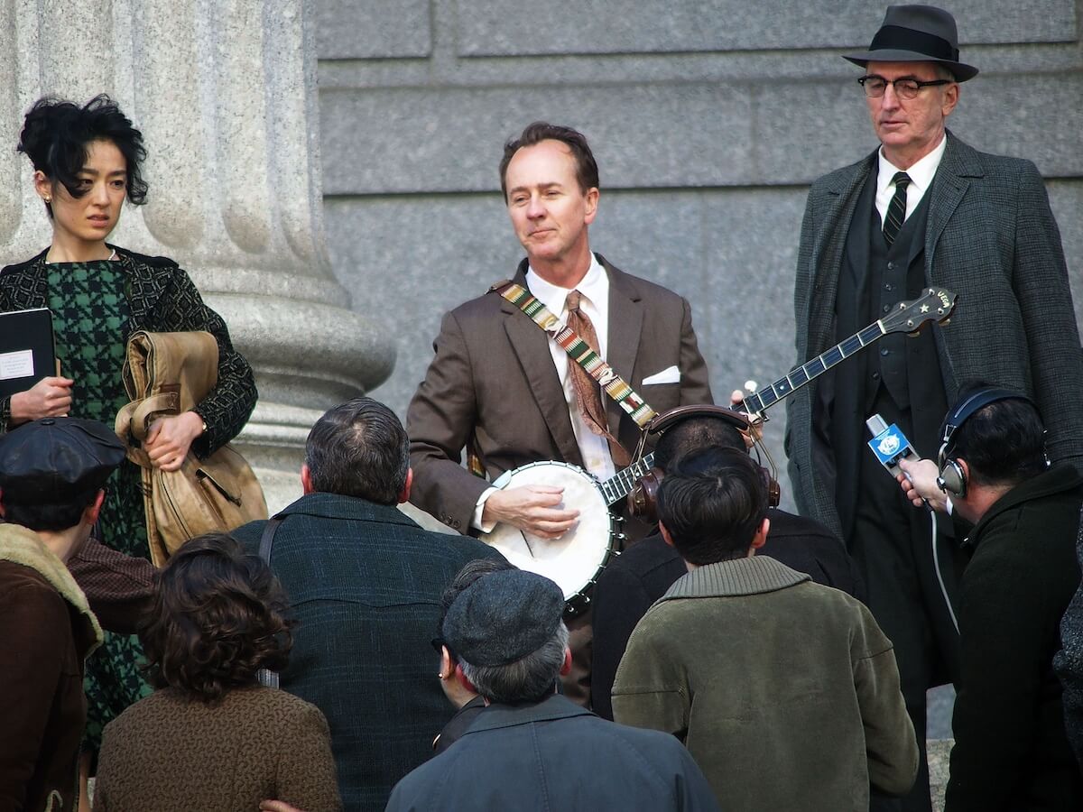 Edward Norton as Pete Seeger holding a banjo and standing on courthouse steps while filming 'A Complete Unknown'