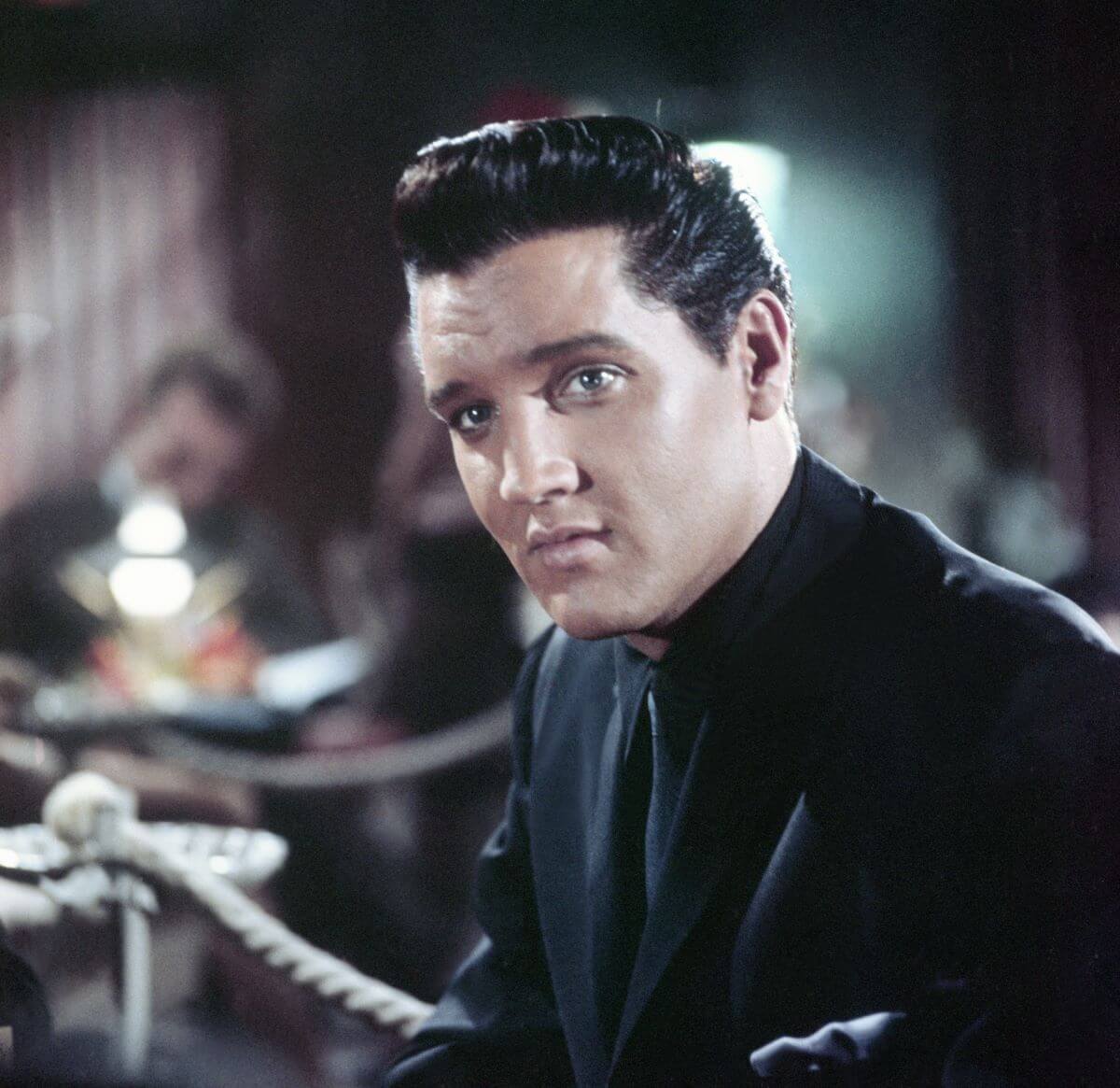 Elvis wears a black suit and sits at a bar.