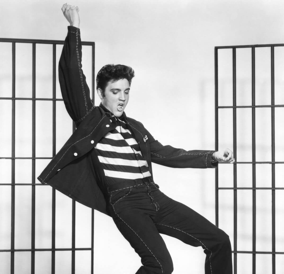 A black and white picture of Elvis wearing a striped shirt and dancing in 'Jailhouse Rock.' He lifts one arm above his head.