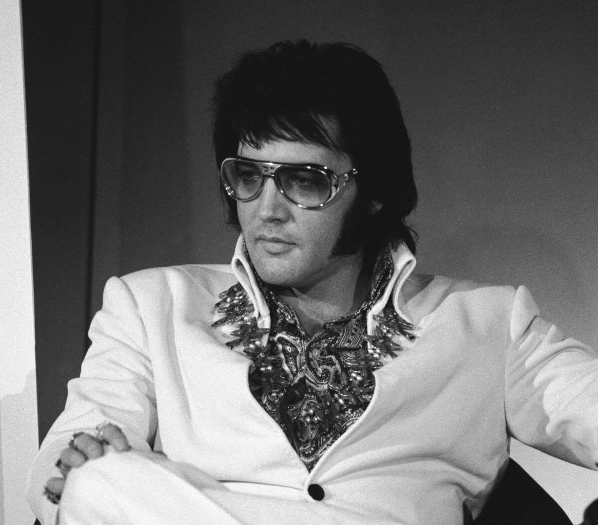 A black and white picture of Elvis Presley wearing a jumpsuit and sunglasses. He sits and rests one hand on his knee.