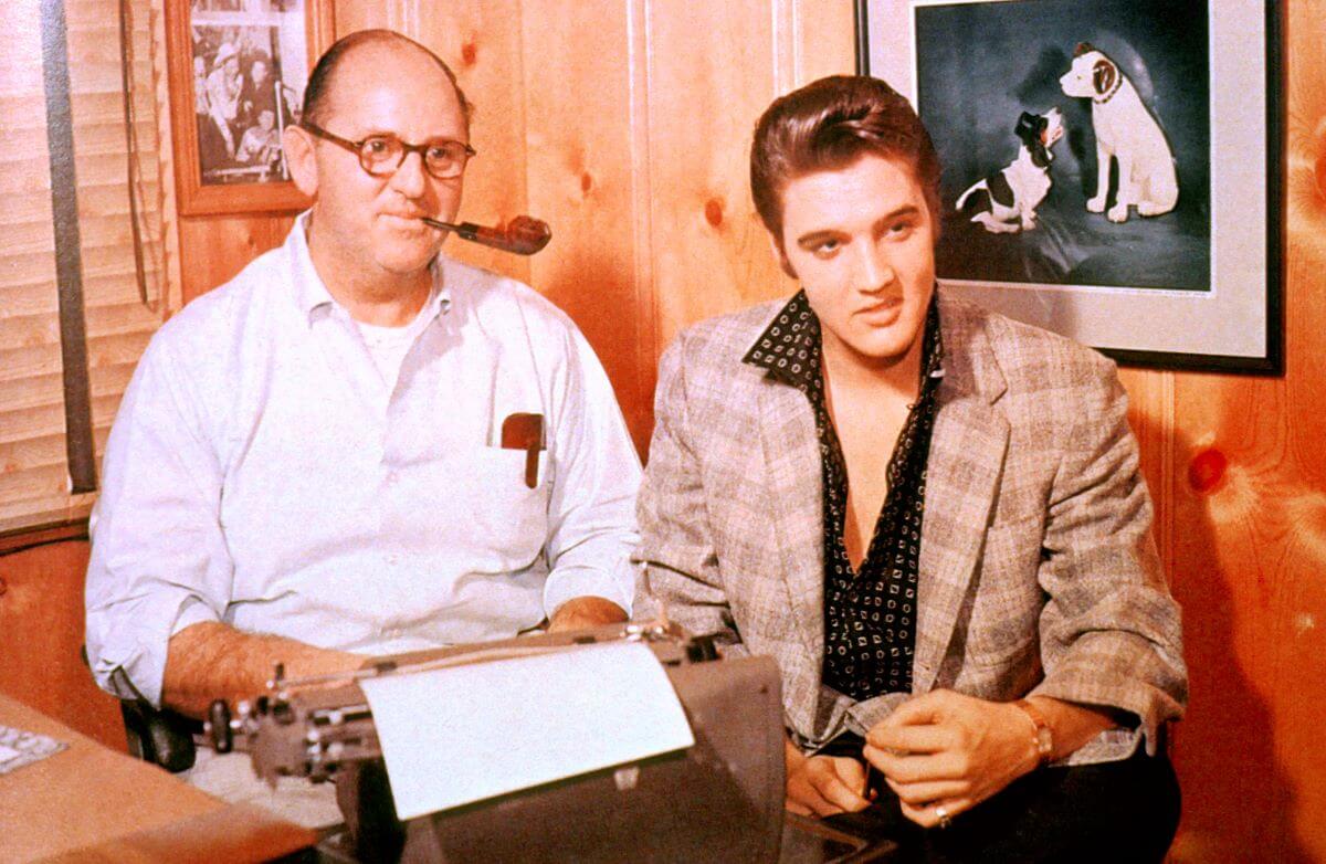 Colonel Tom Parker holds a pipe in his mouth and sits next to Elvis Presley. They sit in front of a wood paneled wall.