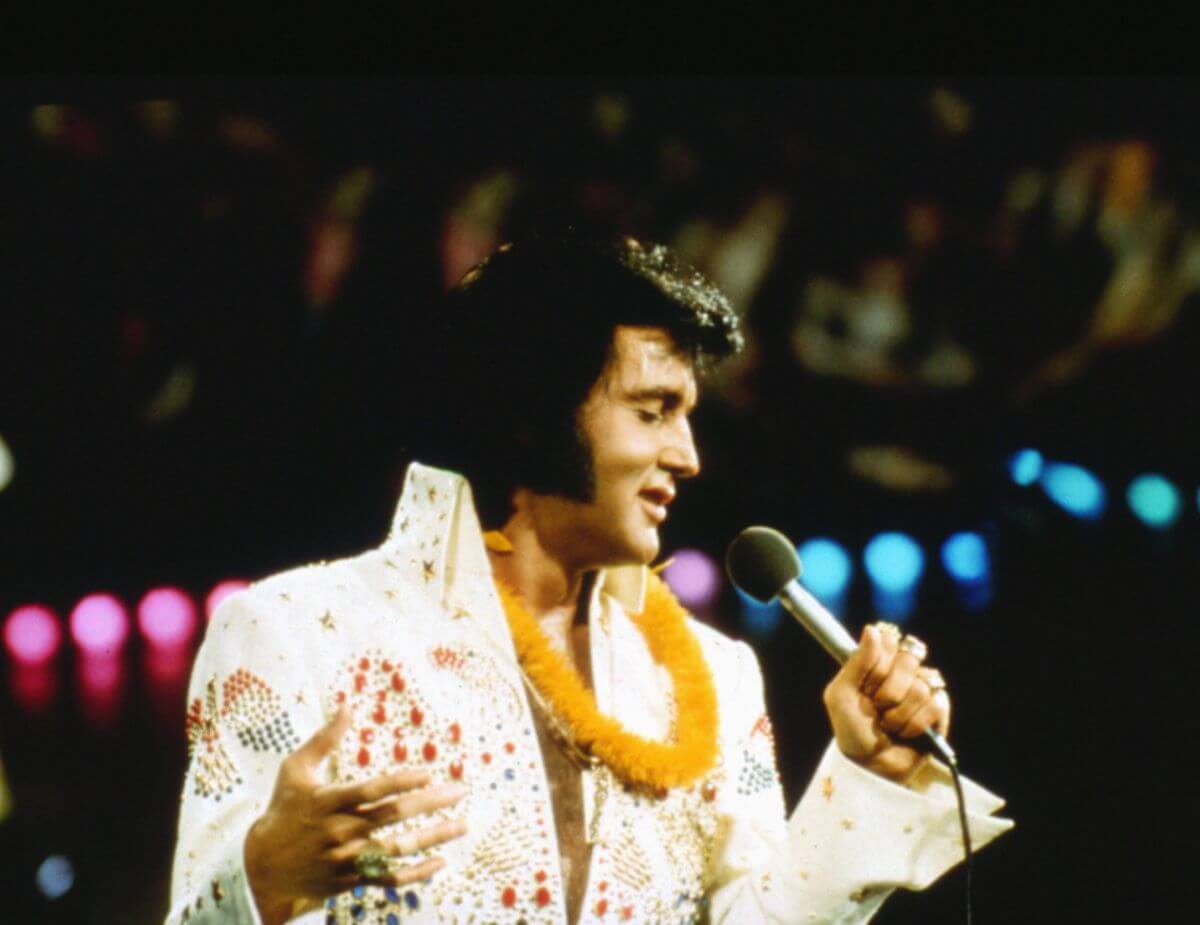 Elvis Presley wears a white jumpsuit and a lei. He speaks into a microphone.
