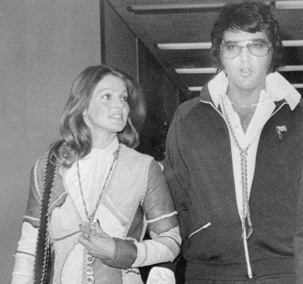 A black and white picture of Priscilla and Elvis Presley walking together. They both wear coats.