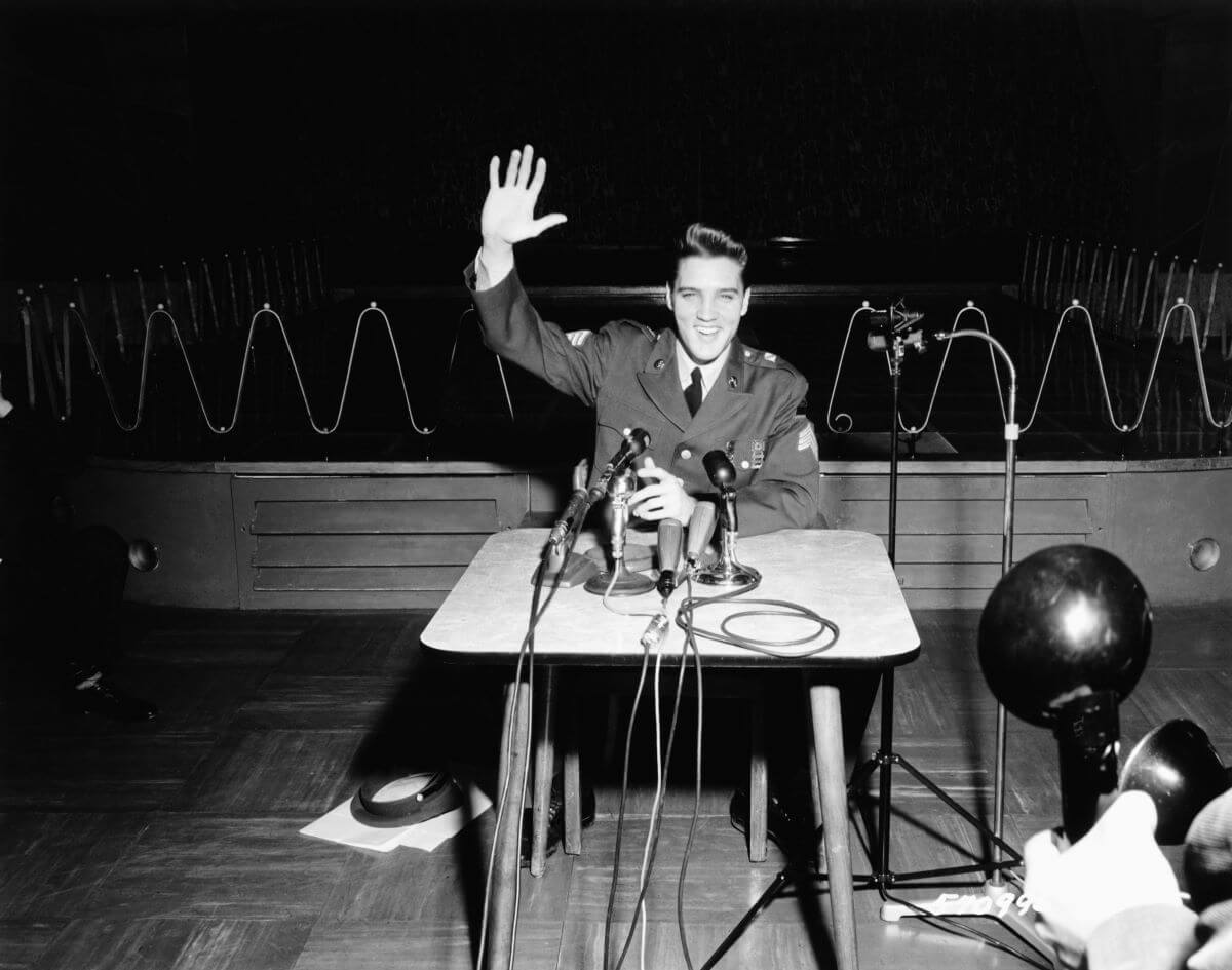 A black and white picture of Elvis in his army uniform and sitting at a desk during a press conference. He waves one arm and smiles.