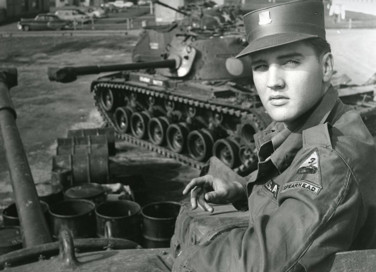 A black and white picture of Elvis in his army uniform and sitting near a tank.