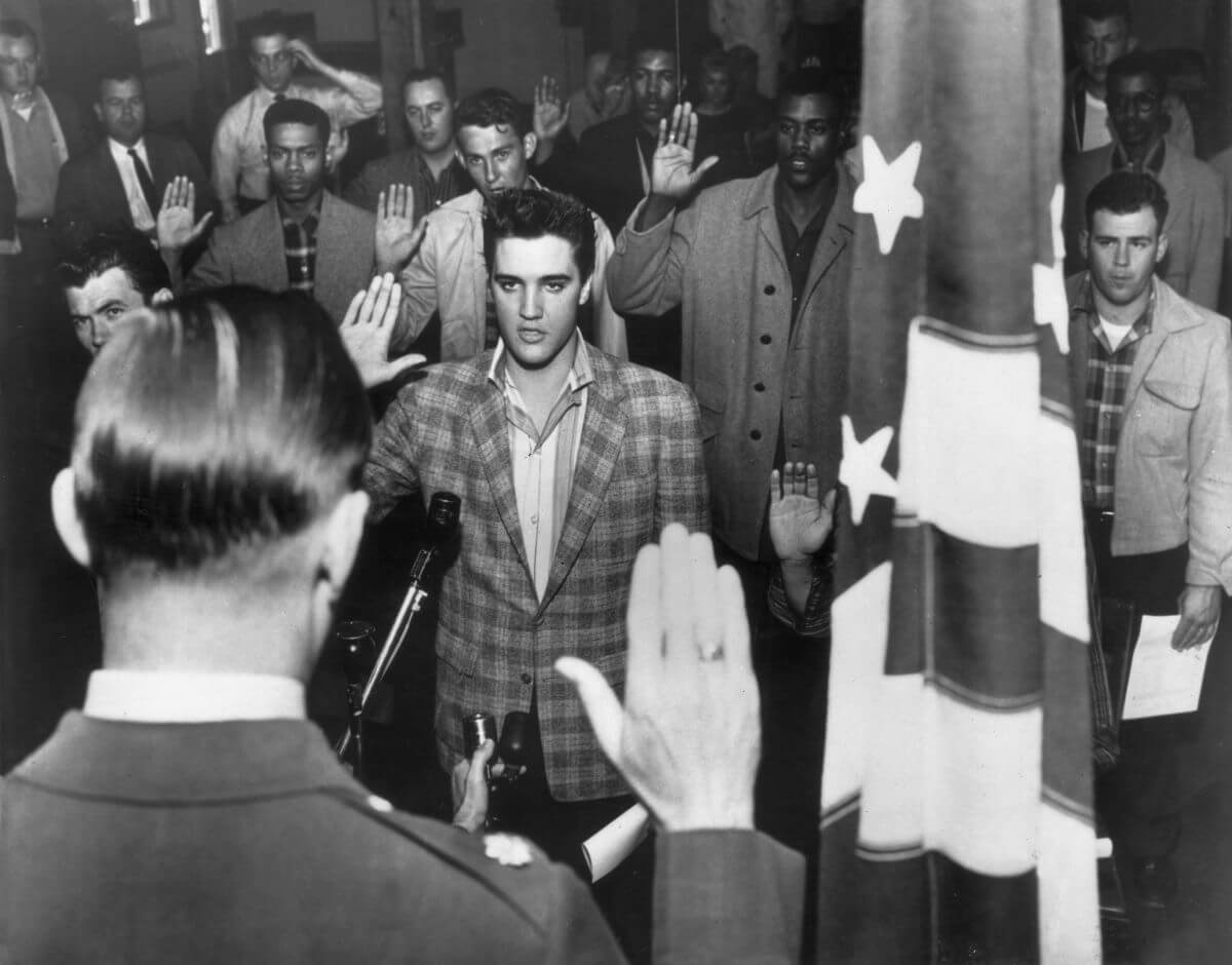 A black and white picture of Elvis raising his hand as he is inducted into the army with other men.