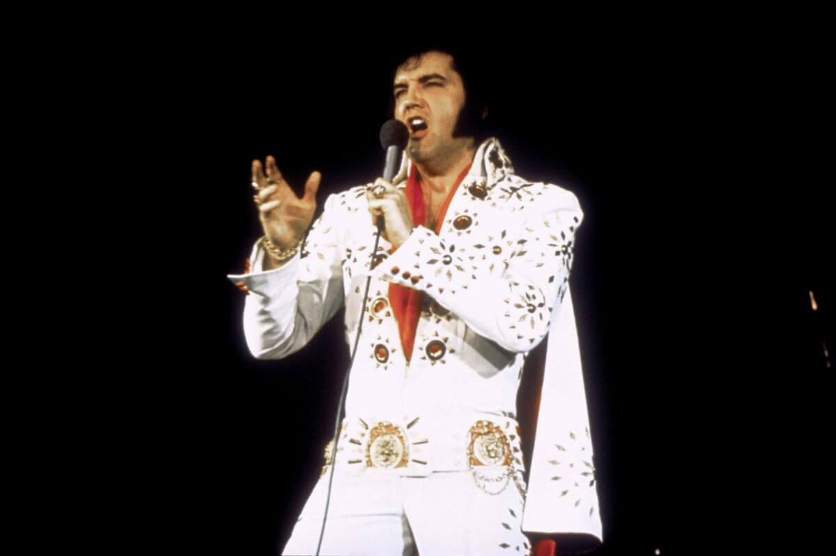 Elvis Presley wears a white jumpsuit and red scarf. He sings into a microphone.