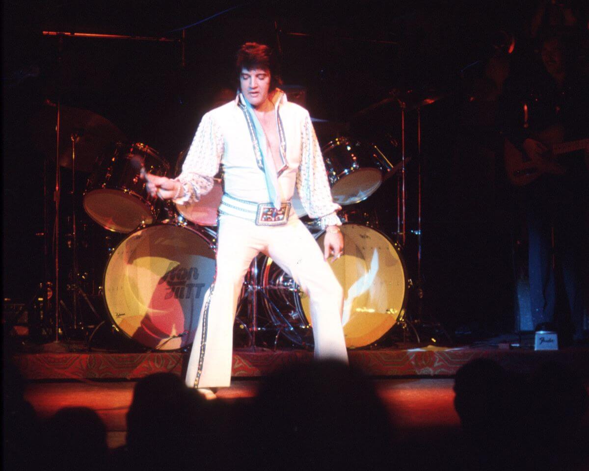Elvis Presley wears a white jumpsuit and holds a microphone on stage.
