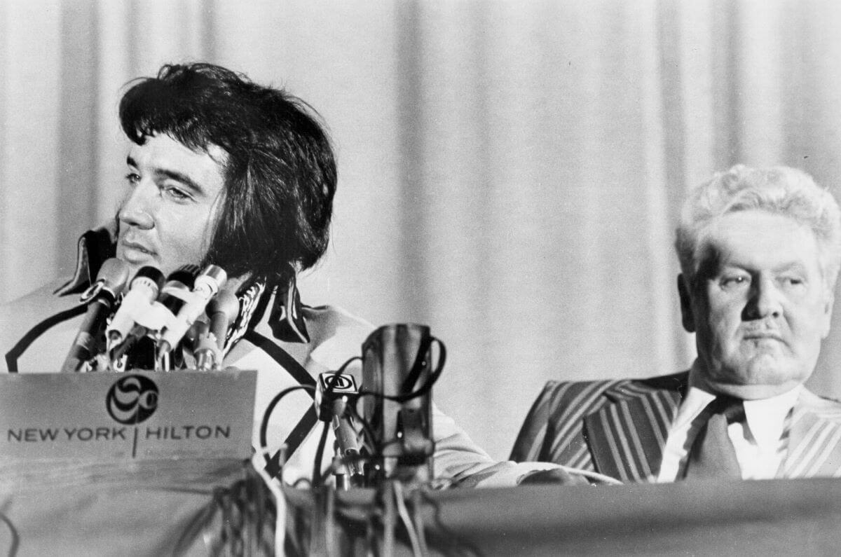 A black and white picture of Elvis sitting in front of a microphone. His father, Vernon, sits next to him.