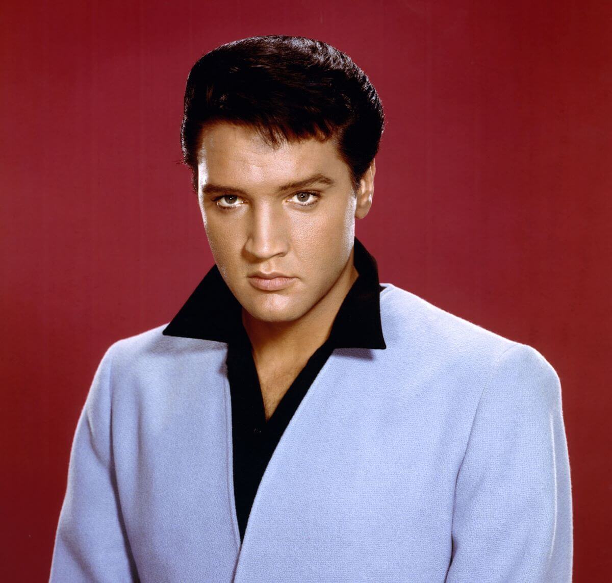 Elvis wears a blue suit and poses in front of a red background.