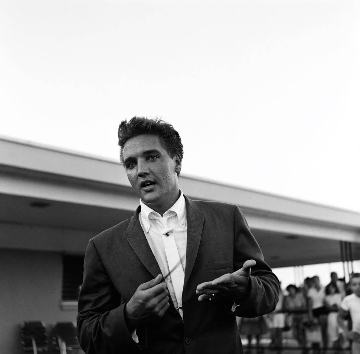 A black and white picture of Elvis Presley standing outside wearing a suit.
