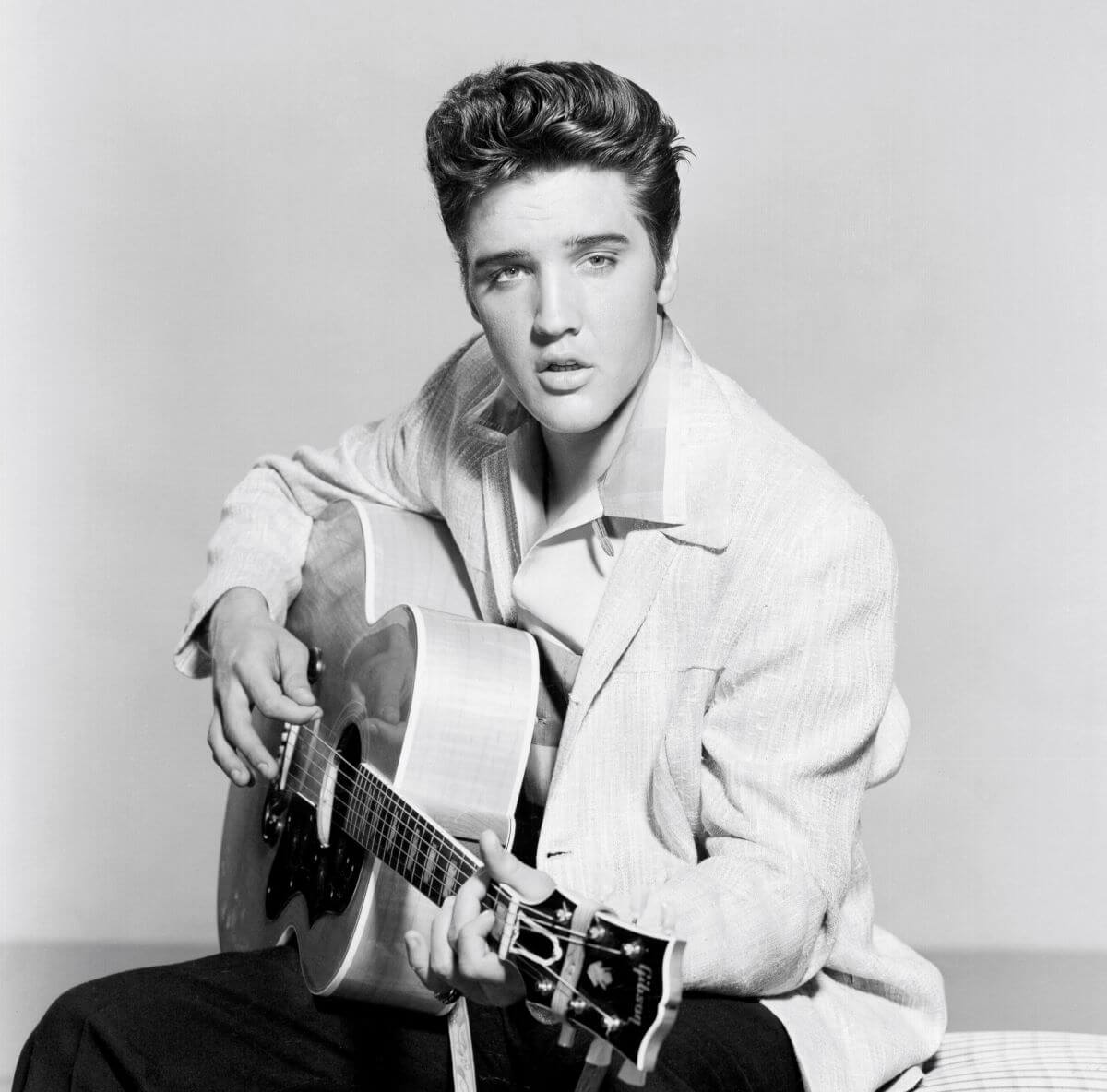 A black and white picture of Elvis sitting with an acoustic guitar.