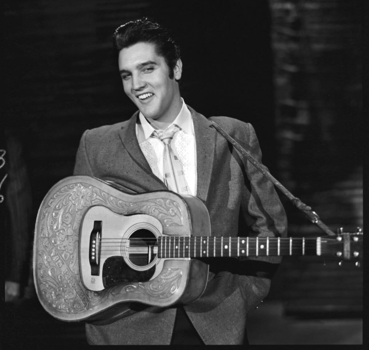 A black and white picture of Elvis wearing a suit and holding an acoustic guitar.