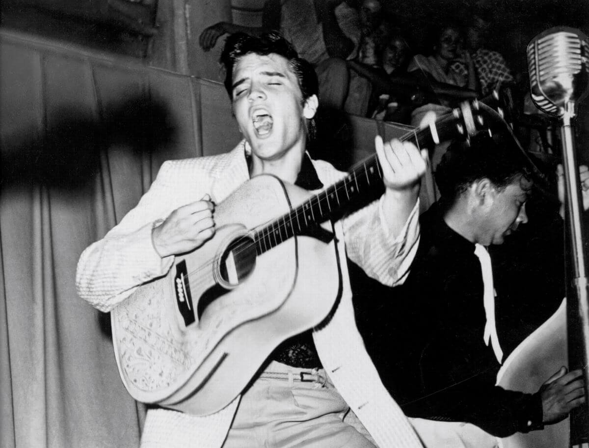 A black and white picture of Elvis strumming an acoustic guitar and singing a song.