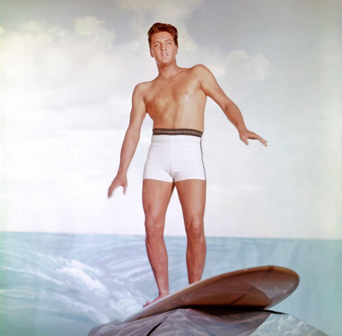 Elvis wears white swim trunks and stands on a surfboard for a publicity shot.