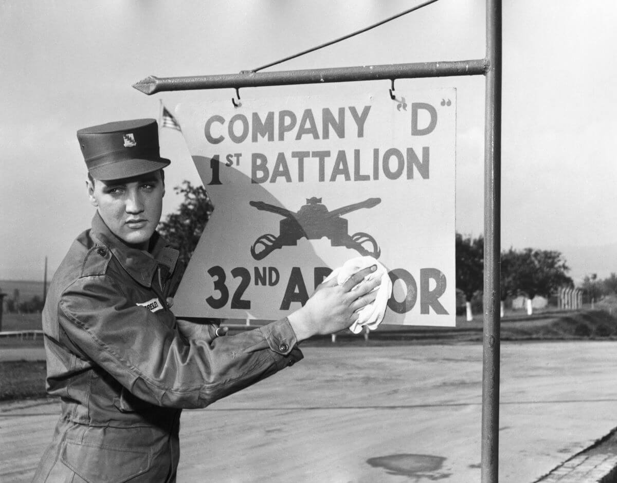 A black and white picture of Elvis wearing his army uniform and cleaning a sign with a rag.