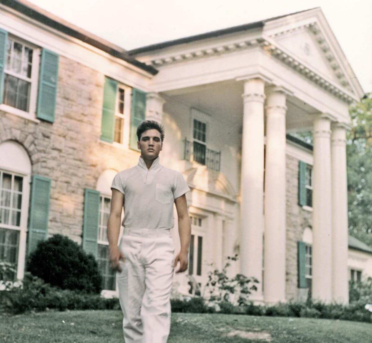 Elvis wears white pants and a white shirt. He walks in front of Graceland.
