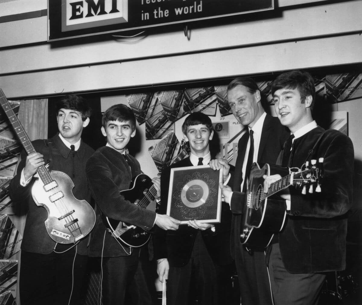A black and white picture of Paul McCartney, George Harrison, Ringo Starr, and John Lennon of The Beatles standing with producer George Martin. McCartney, Harrison, and Lennon hold guitars and Starr holds a framed record.