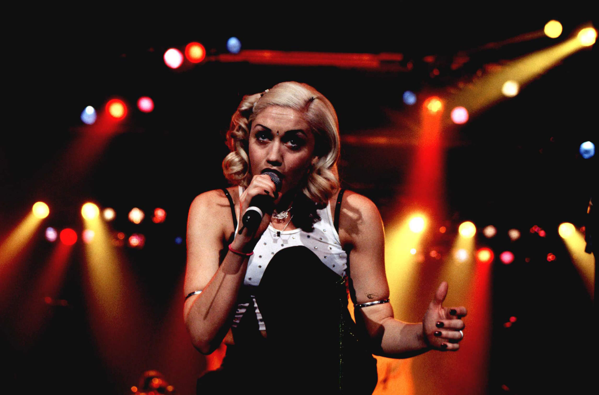 Gwen Stefani kneeling on stage while holding a microphone to her mouth in 1997