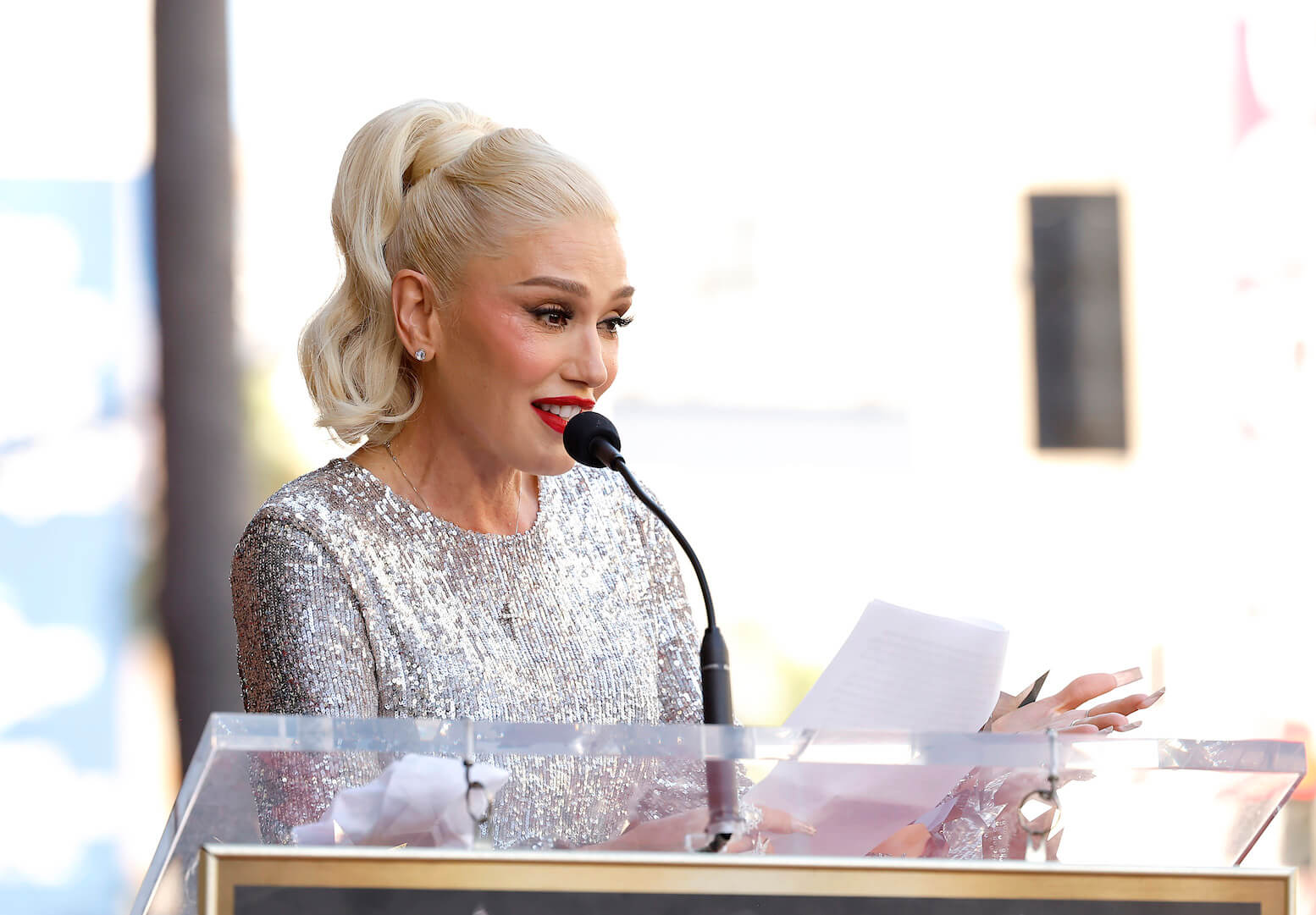 Gwen Stefani speaking into a microphone on a podium while wearing a sparkly silver dress on the Hollywood Walk of Fame