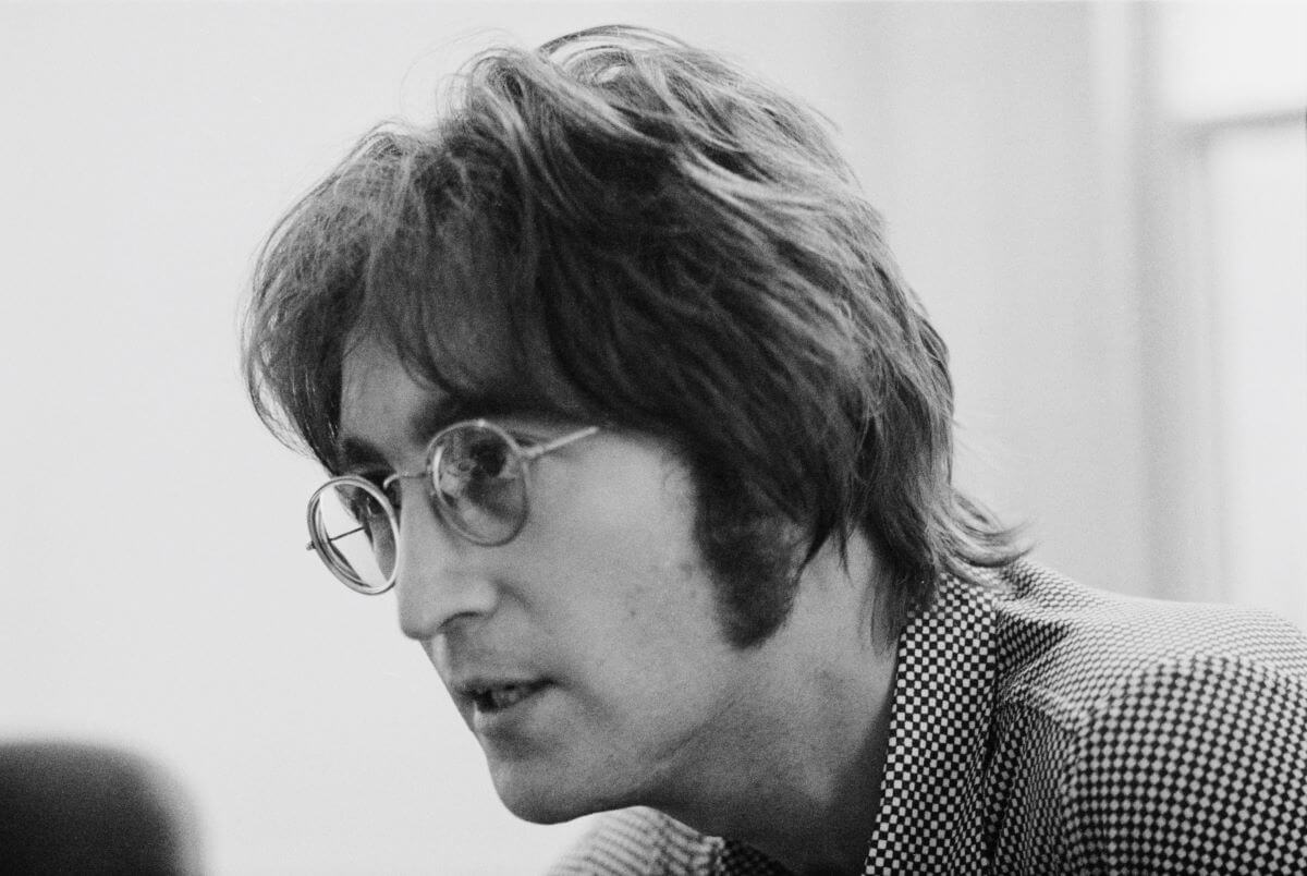 A black and white picture of John Lennon wearing glasses and a checkered shirt. He sits near a window.