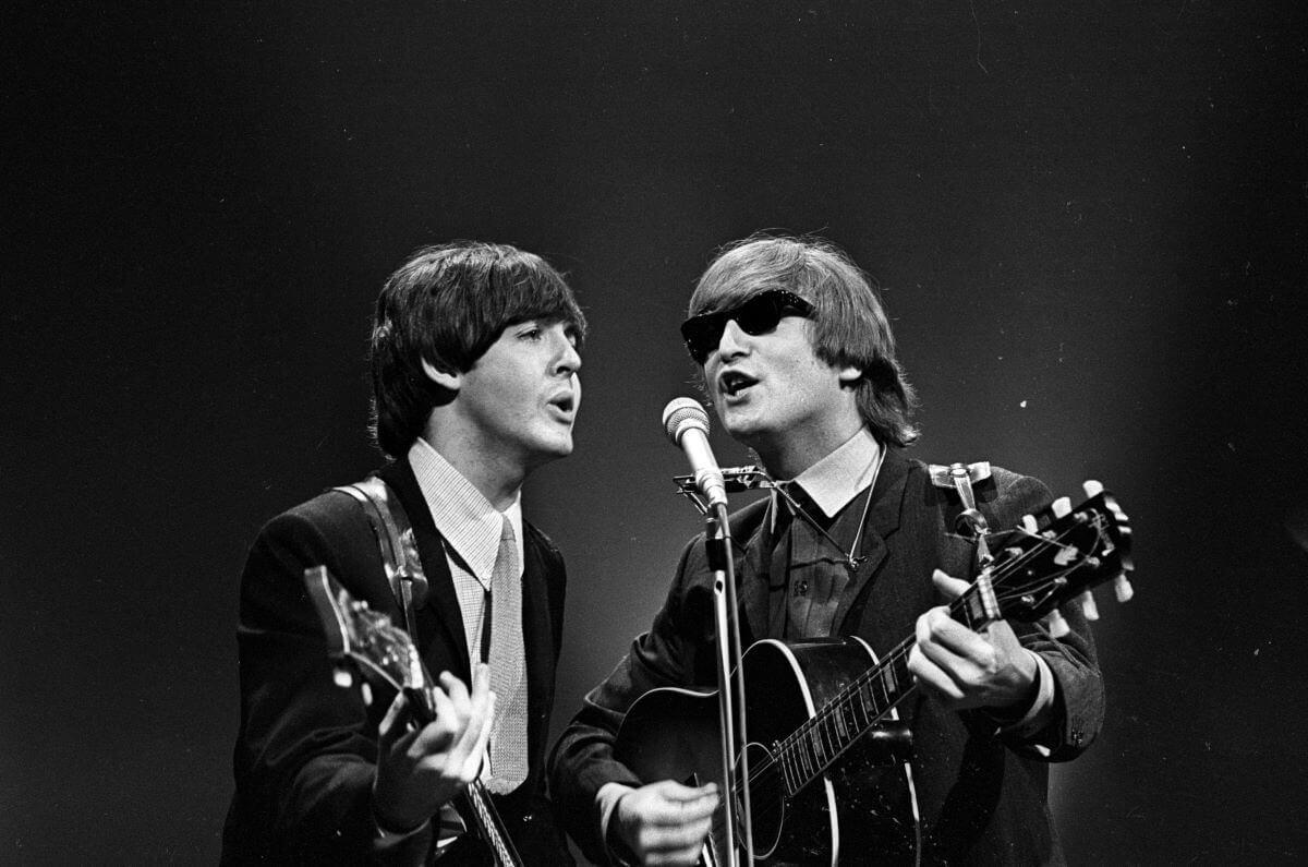 A black and white picture of Paul McCartney and John Lennon playing guitars and singing into the same microphone. Lennon wears sunglasses.