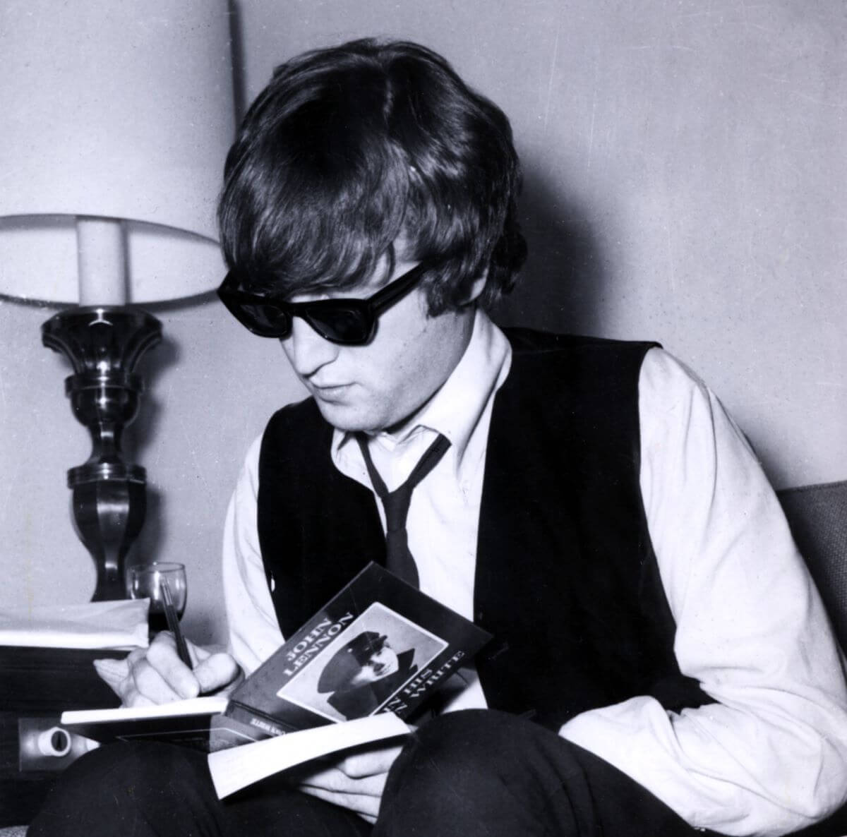 A black and white picture of John Lennon wearing sunglasses and sitting while he signs one of his books.