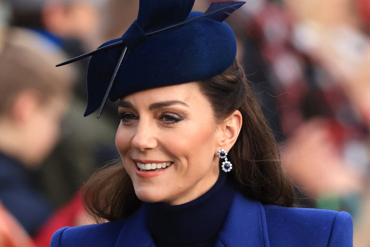 Kate Middleton, who is pictured in a paparazzi photo from March 2024, smiles wearing a blue coat and hat on Christmas Day 2023