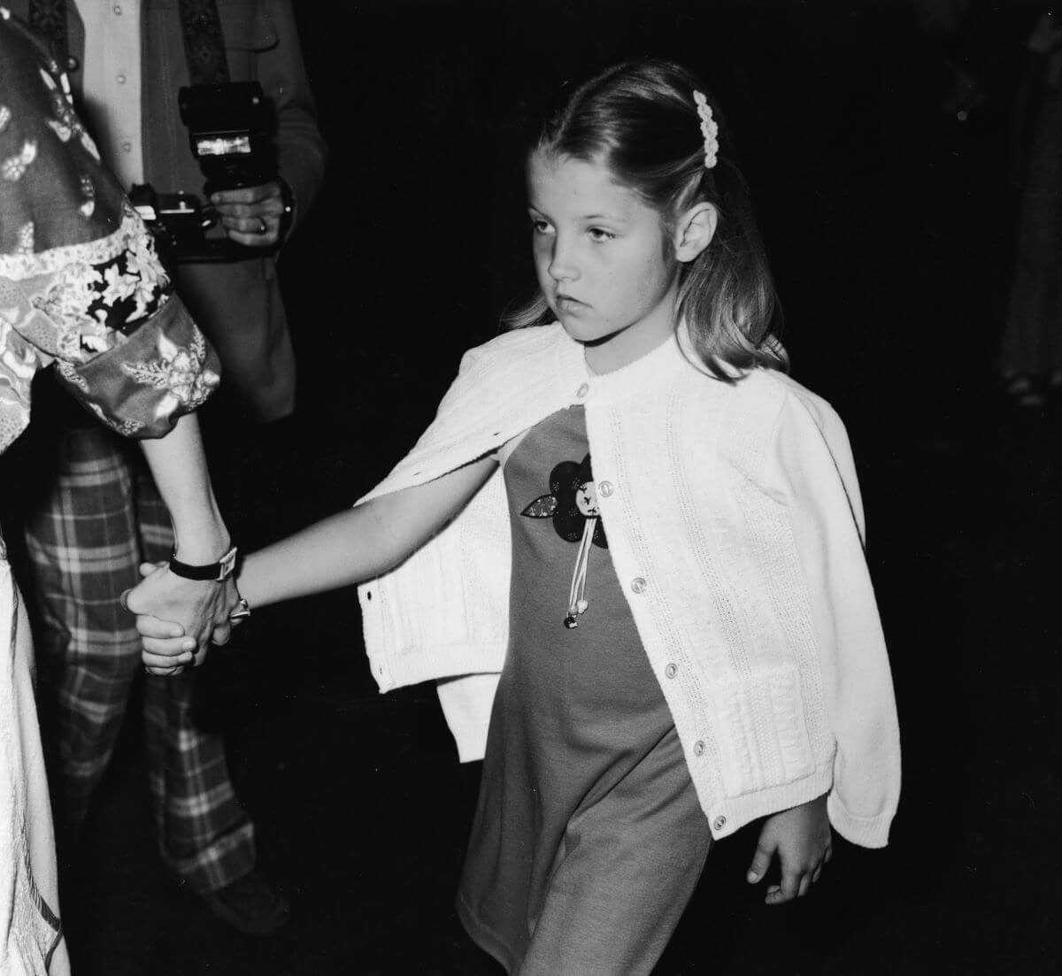 A black and white picture of Lisa Marie Presley wearing a cardigan and holding someone out of frame's hand.