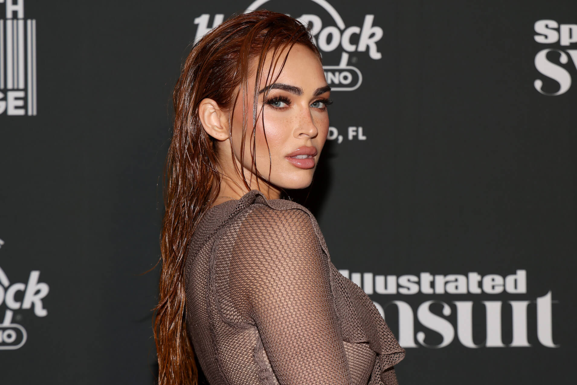 Megan Fox at a Sports Illustrated event looking over her shoulder. Her hair is slicked and she's wearing a mesh sleeved dress