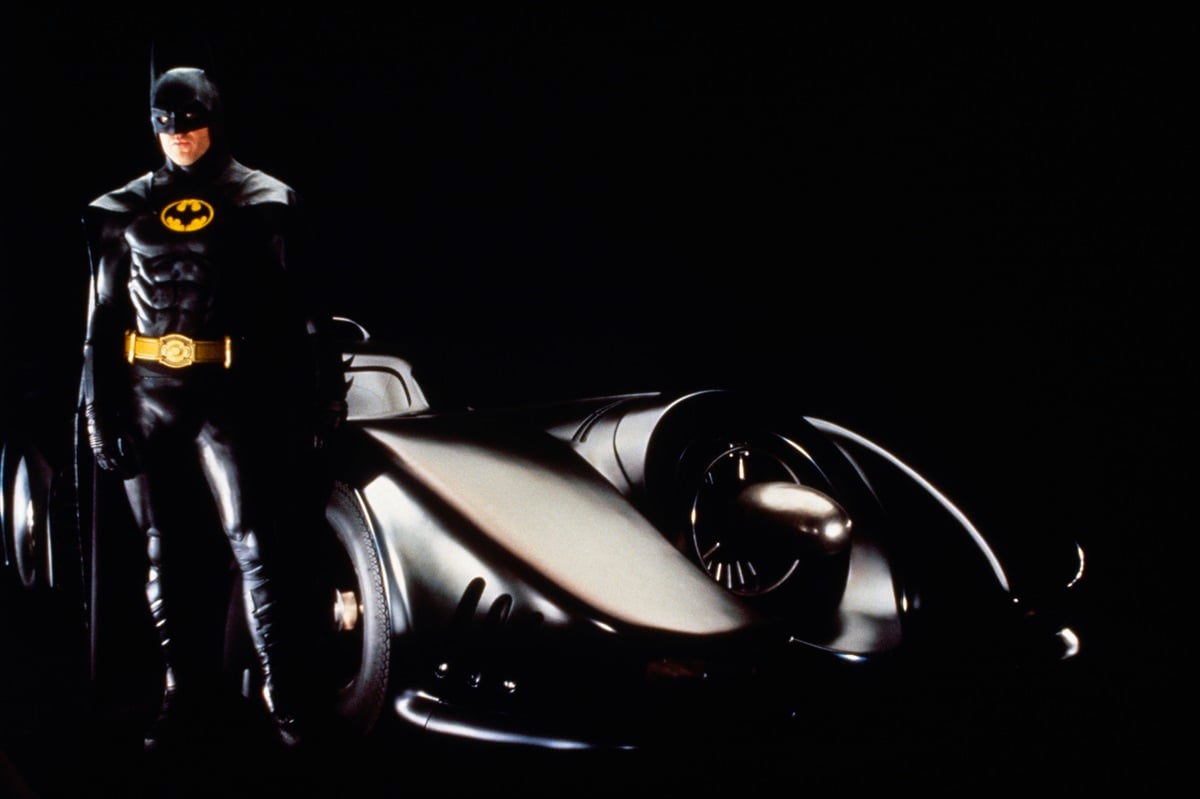 Michael Keaton posing in a Batman costume next to the Batmobile.