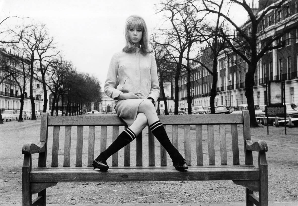 A black and white picture of Pattie Boyd sitting on top of a bench with her legs crossed.