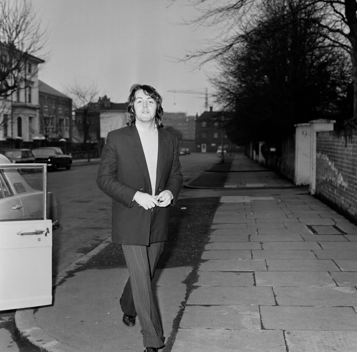 A black and white picture of Paul McCartney walking next to a car.