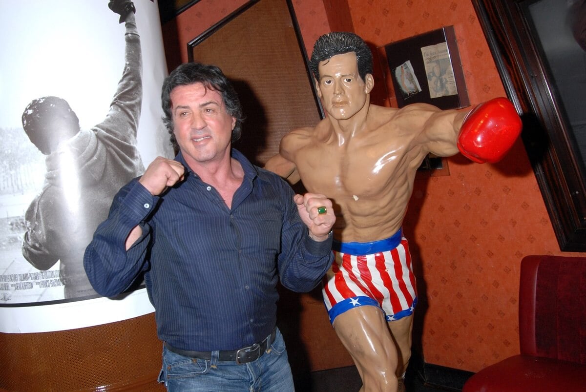 Sylvester Stallone posing at Planet Hollywood next to a Rocky statue.