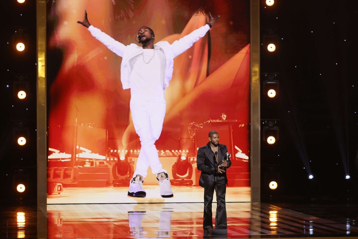 Usher wears a black suit and stands onstage at the NAACP Awards. The screen behind him shows a large image of himself wearing white.