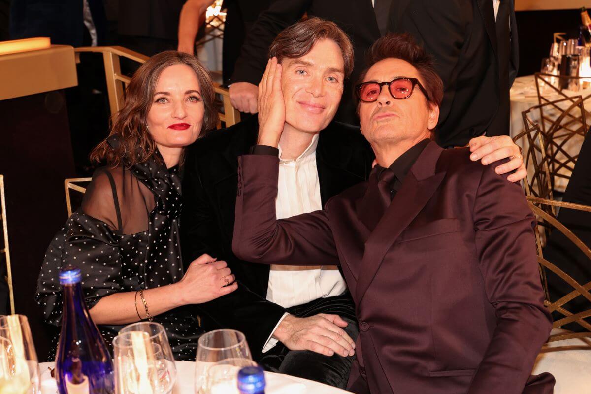 Yvonne McGuinness, Cillian Murphy, and Robert Downey Jr. sit at a table together. McGuinness holds Murphy's arm and Downey Jr. touches his hand to Murphy's face.