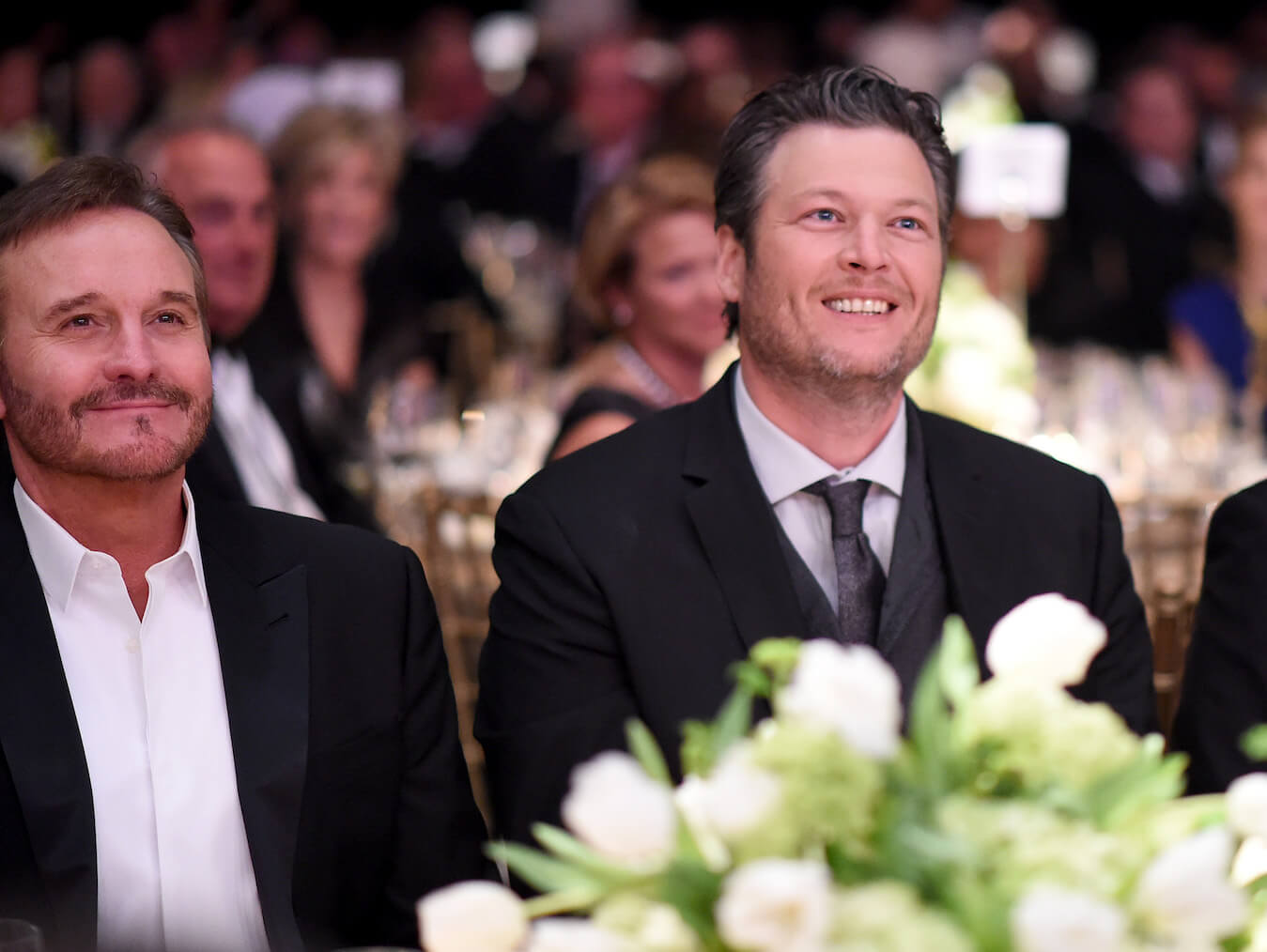 Narvel Blackstock sitting at a table next to Blake Shelton. They are both wearing suits and smiling while looking up at something in front of them.