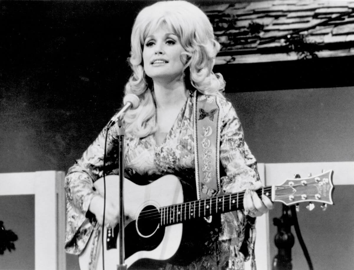 A black and white picture of Dolly Parton standing in front of a microphone and holding a guitar.