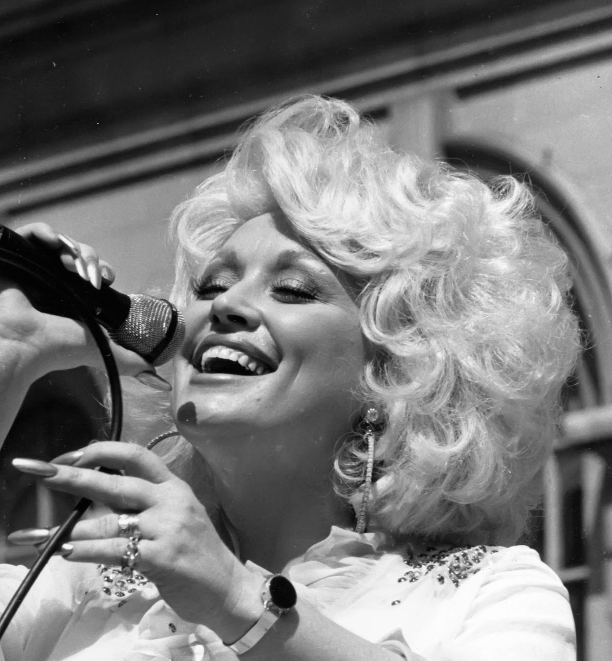 A black and white picture of Dolly Parton singing into a microphone. She holds the wire as she smiles.