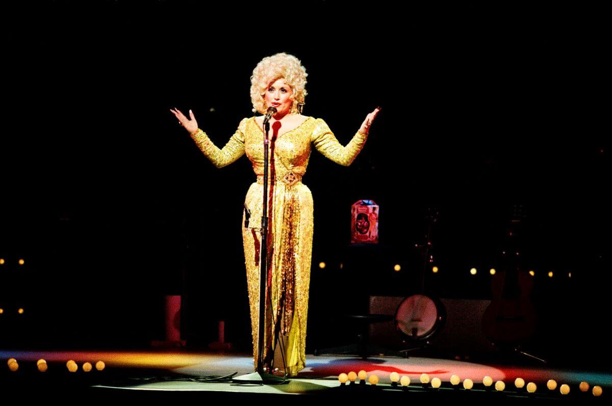 Dolly Parton stands in front of a microphone and lifts her arms.