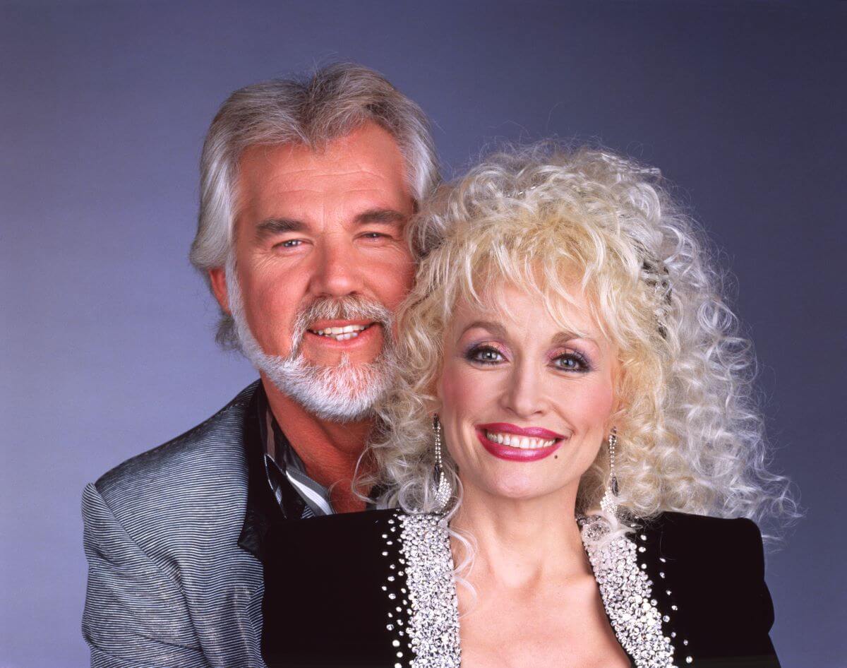 Kenny Rogers stands behind Dolly Parton with his arms around her. He wears a silver jacket and she wears a black jacket.