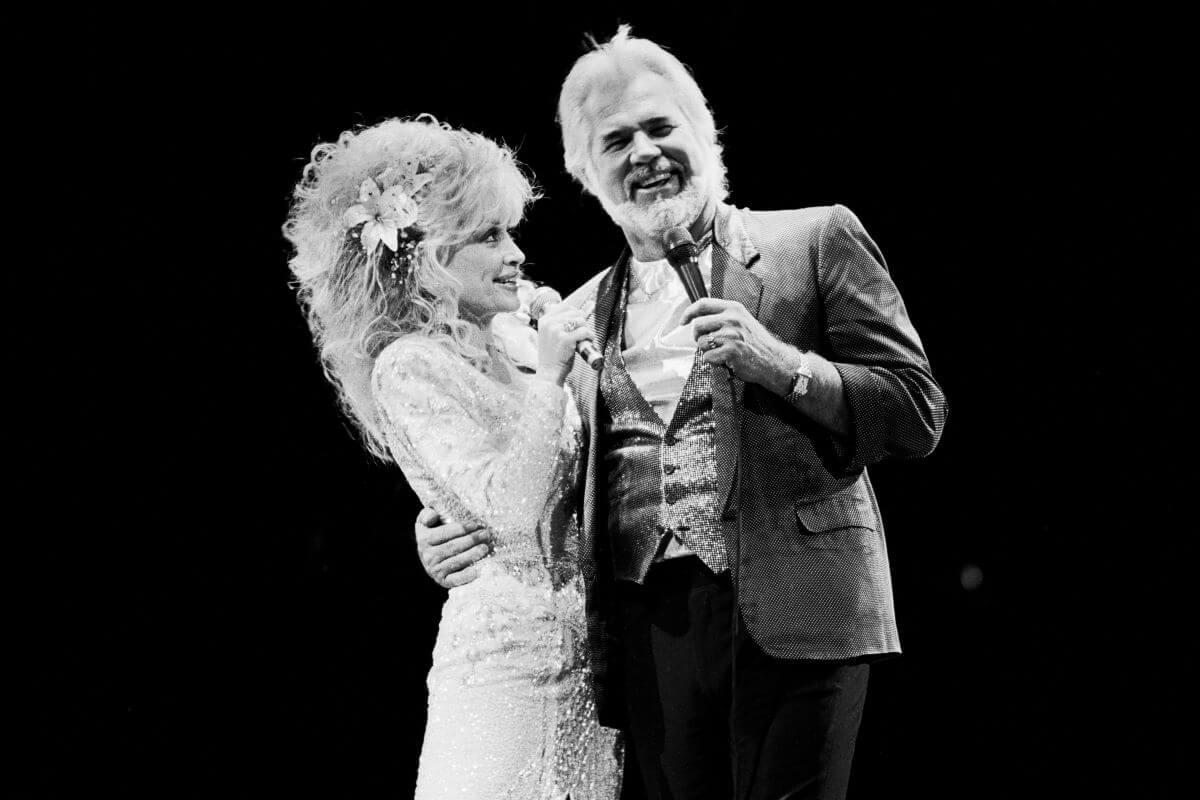 A black and white picture of Kenny Rogers standing with his arm around Dolly Parton. She looks up at him. They both hold microphones.