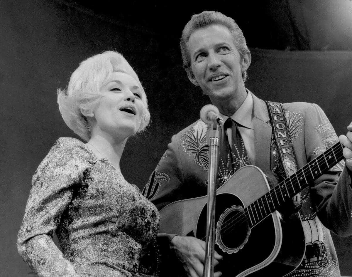 A black and white picture of Dolly Parton and Porter Wagoner singing into a microphone. He holds an acoustic guitar.
